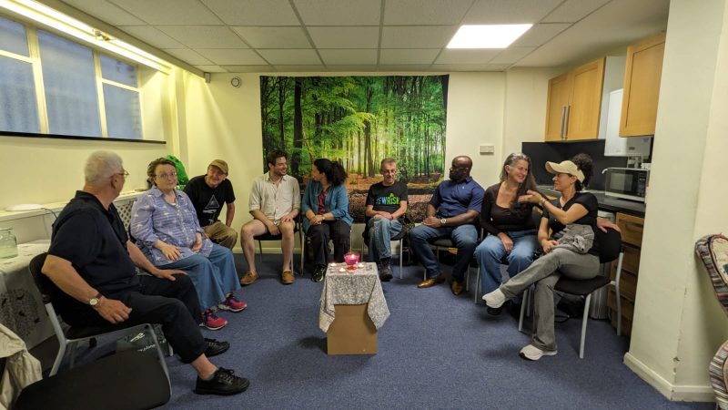 A gathering of happy people, sitting in a circle, all in discussion with their neighbour. There is a huge poster of a rainforest in the background, on the far wall, the rug is blue, the walls are a warm yellow. In the middle stands a table with tablecloth on it: on top, you can see a lit candle in a red, glass candle holder, as well as three lit tealights surrounding it. The atmosphere is warm and friendly.